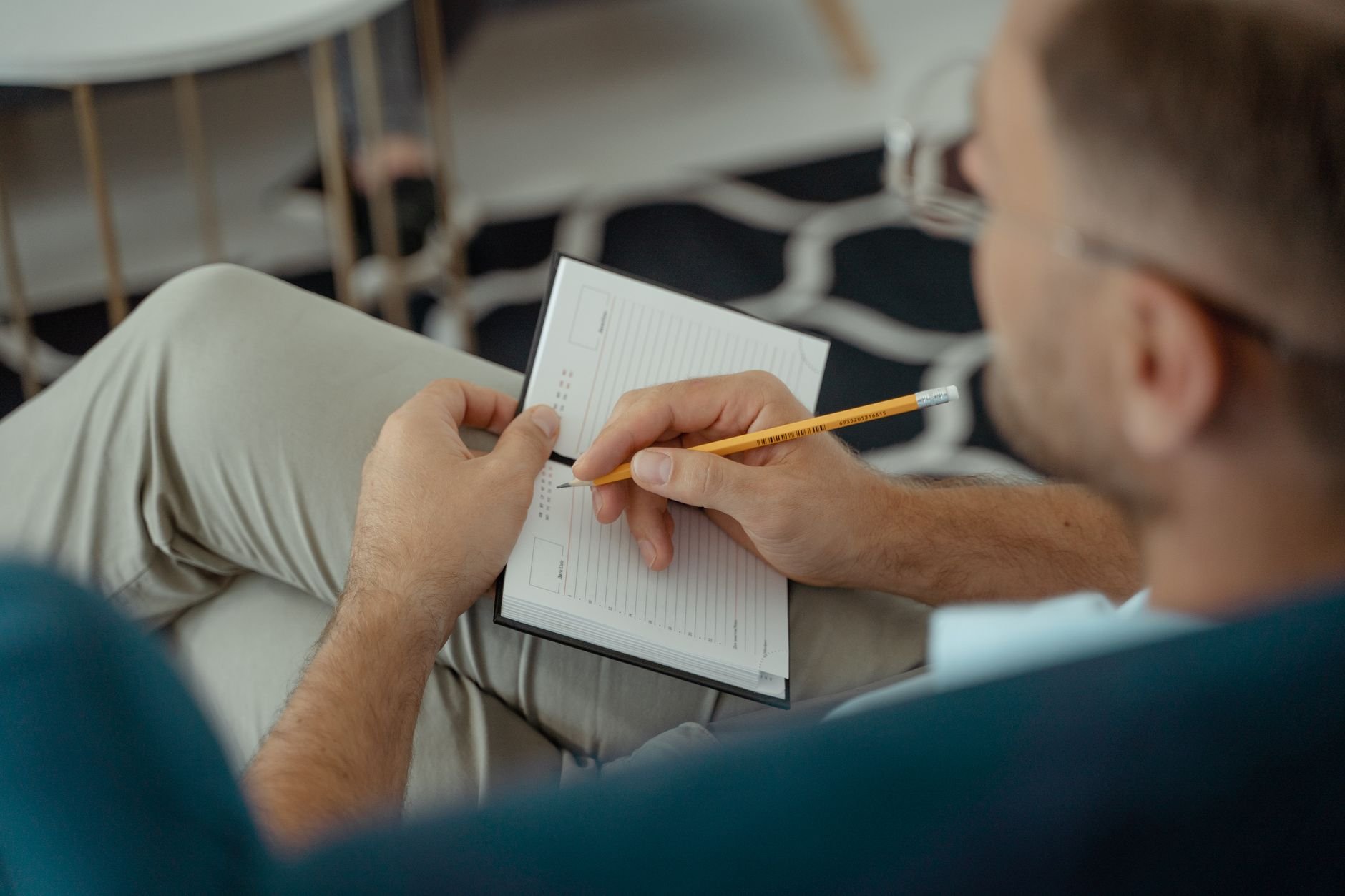 An adult man writing with a pencil in a notebook while seated indoors.
